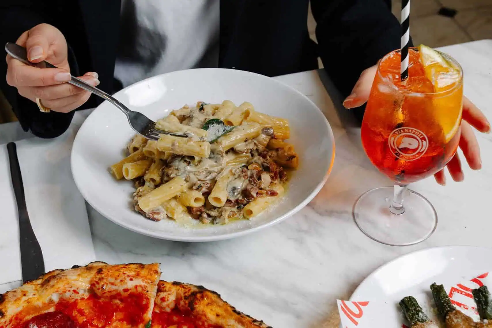A woman enjoys date night at Via Napoli Pizzeria with Neapolitan pepperoni pizza, creamy pasta, and an Aperol Spritz on a marble table.