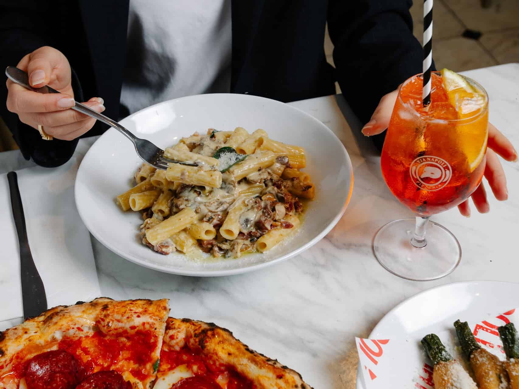 A woman enjoys date night at Via Napoli Pizzeria with Neapolitan pepperoni pizza, creamy pasta, and an Aperol Spritz on a marble table.