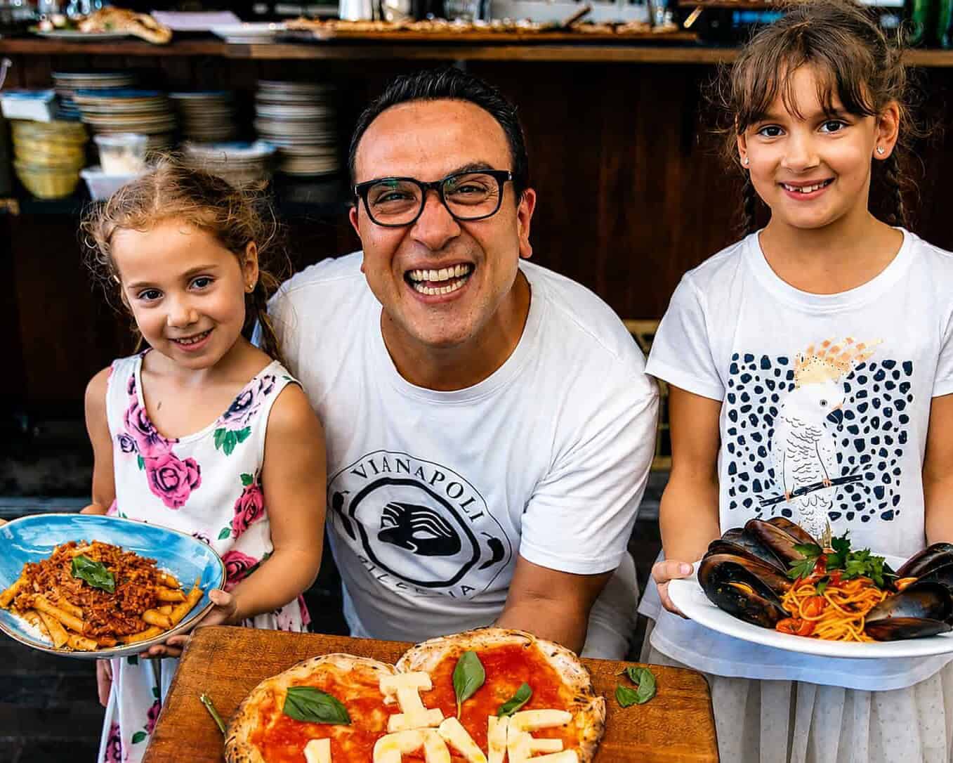 Luigi Espositio smiling with two children holding plates of Italian pasta, with a wood-fired pizza on a board in an Italian restaurant kitchen.