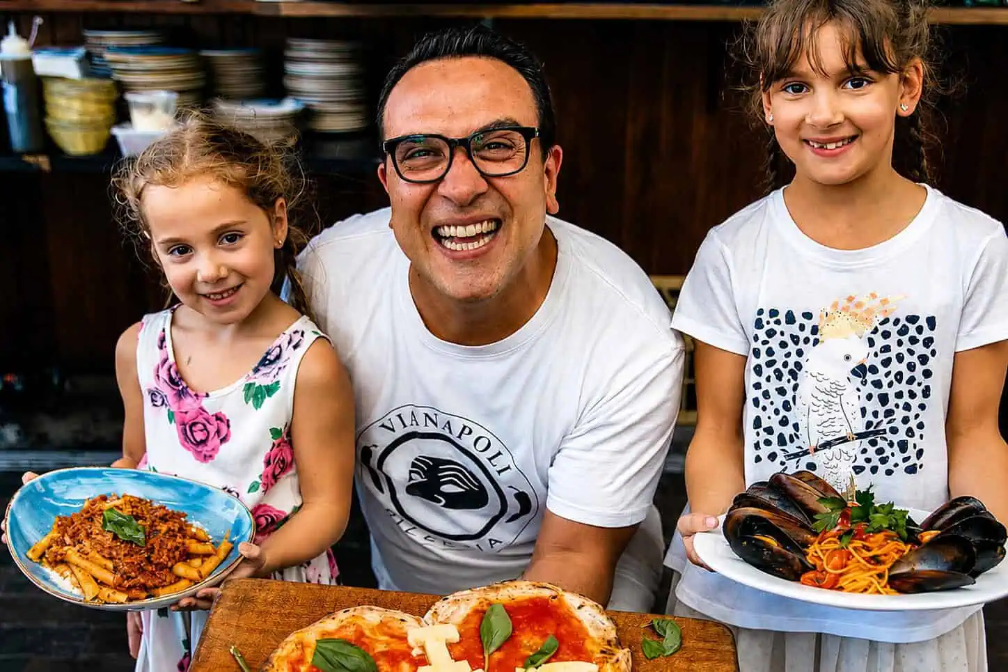 Luigi Espositio smiling with two children holding plates of Italian pasta, with a wood-fired pizza on a board in an Italian restaurant kitchen.