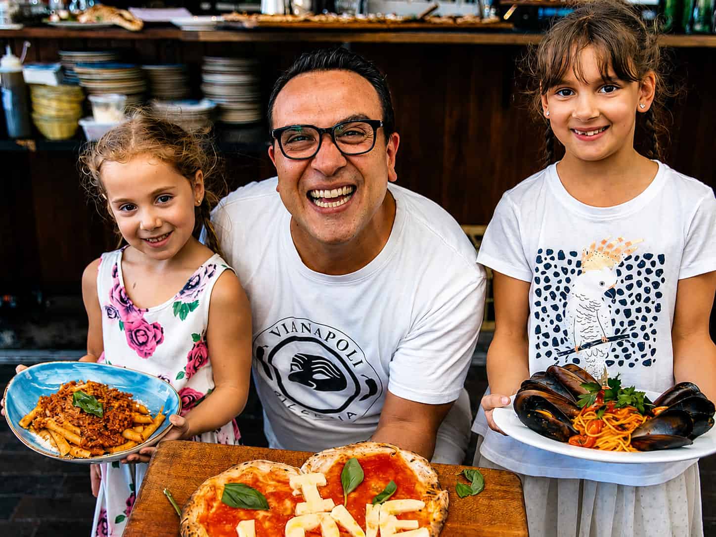 Luigi Espositio smiling with two children holding plates of Italian pasta, with a wood-fired pizza on a board in an Italian restaurant kitchen.