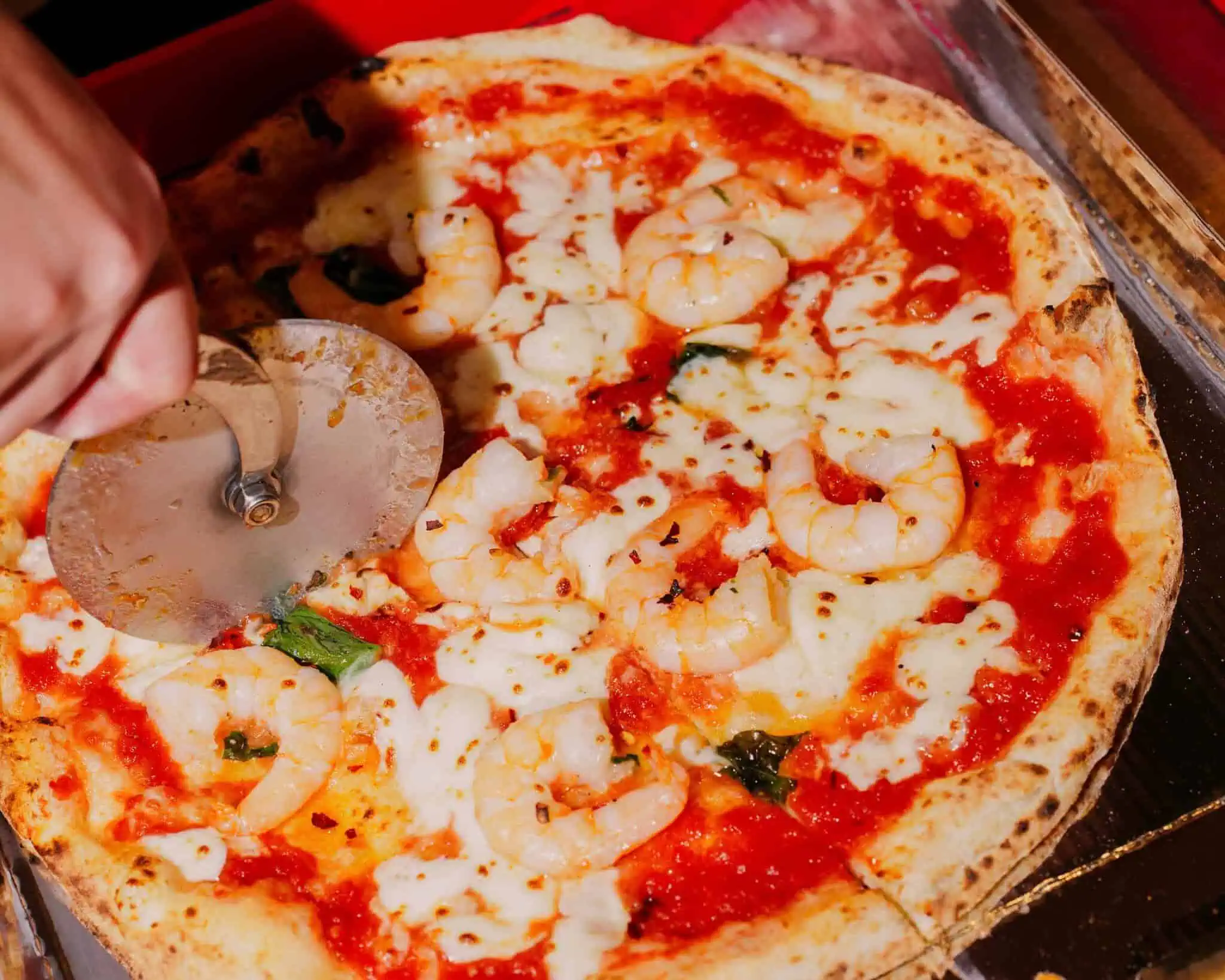 Hand slicing a fresh pizza with melted mozzarella and tomato sauce at Via Napoli Pizzeria
