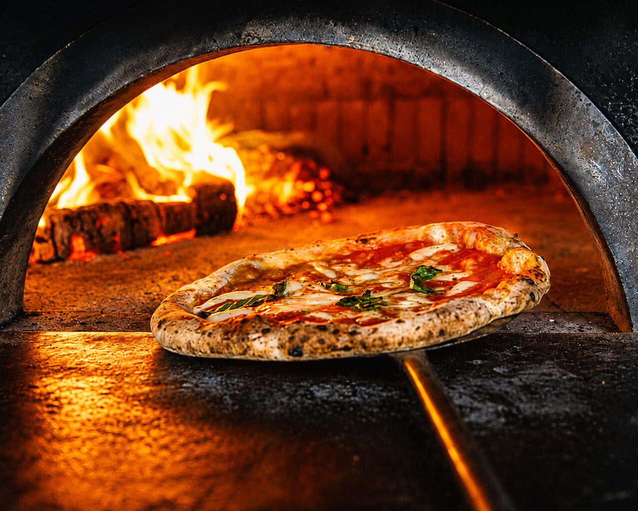 Neapolitan pizza being removed from a wood-fired oven with flames in the background at Via Napoli.
