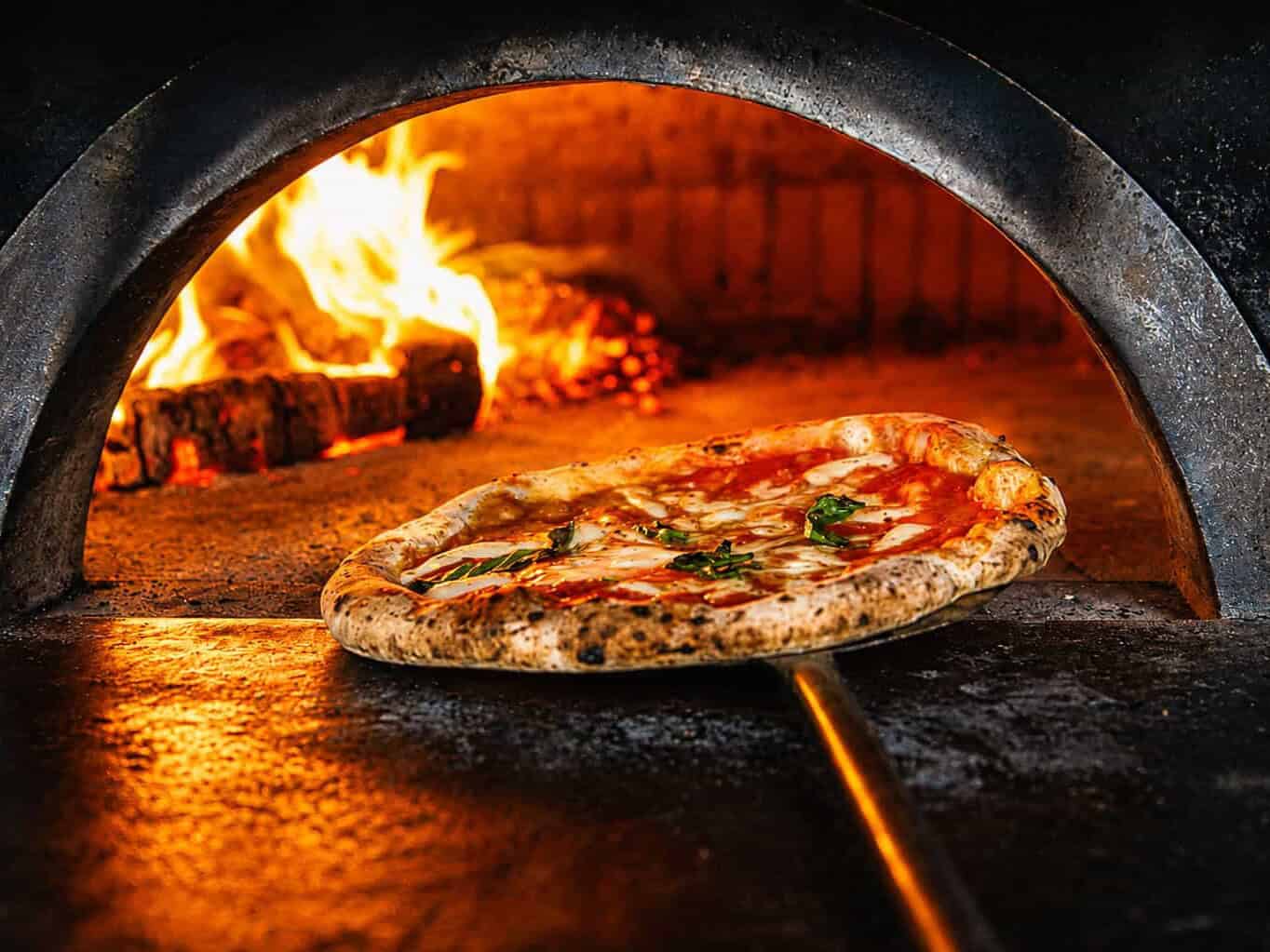 Neapolitan pizza being removed from a wood-fired oven with flames in the background at Via Napoli.