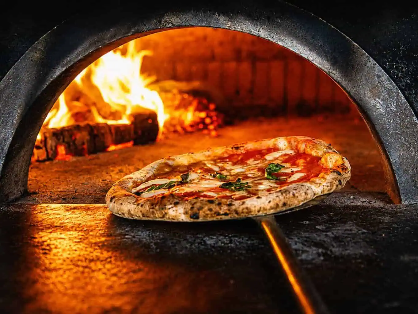 Neapolitan pizza being removed from a wood-fired oven with flames in the background at Via Napoli.