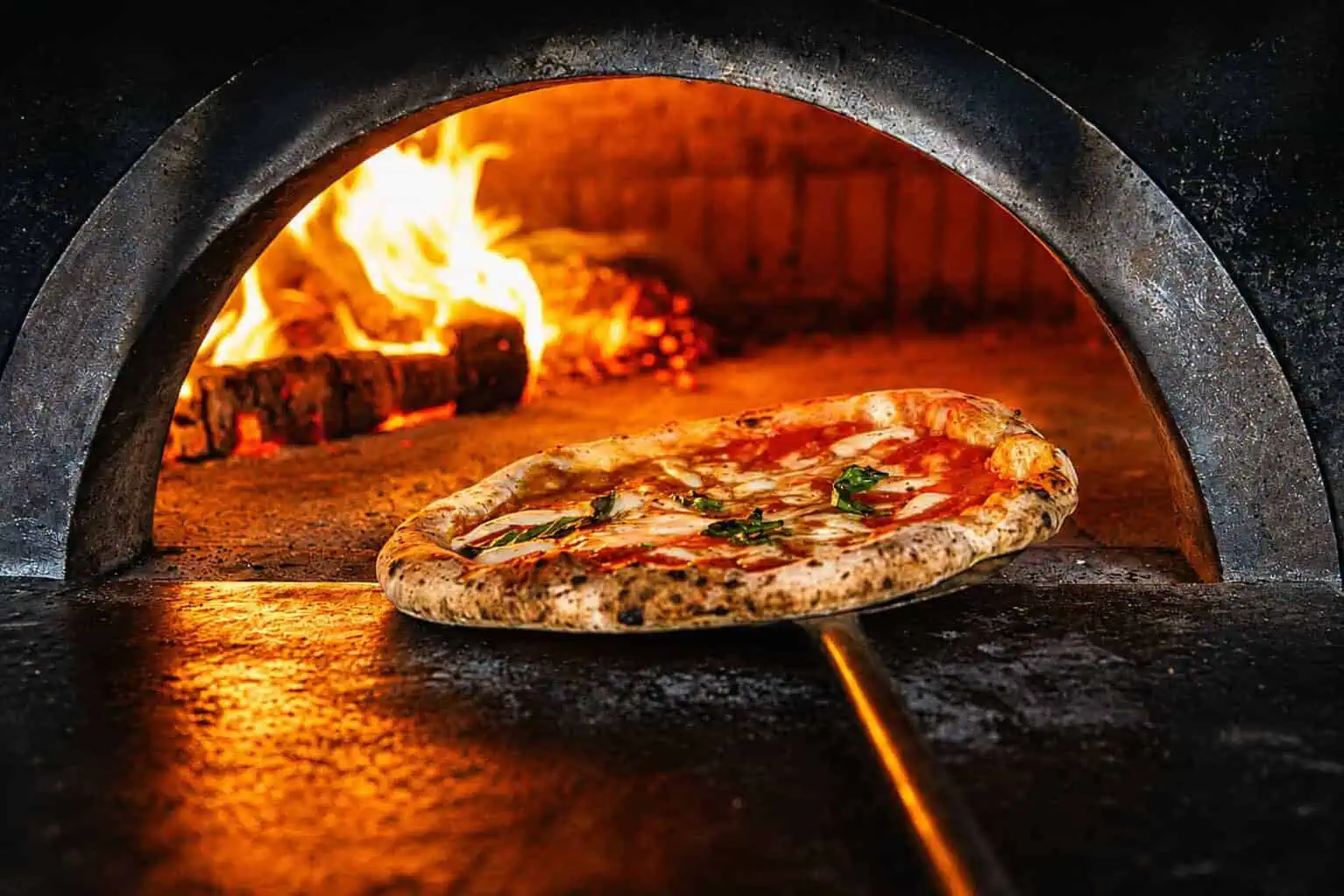 Neapolitan pizza being removed from a wood-fired oven with flames in the background at Via Napoli.