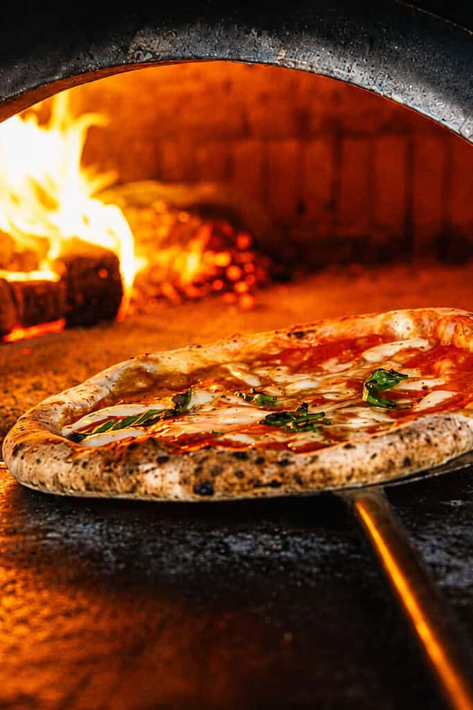 Neapolitan pizza being removed from a wood-fired oven with flames in the background at Via Napoli.