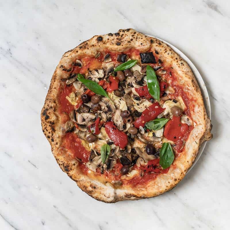 Vegan pizza topped with coconut cheese, San Marzano tomato, roasted capsicum, mushrooms, eggplant, black olives, and fresh basil