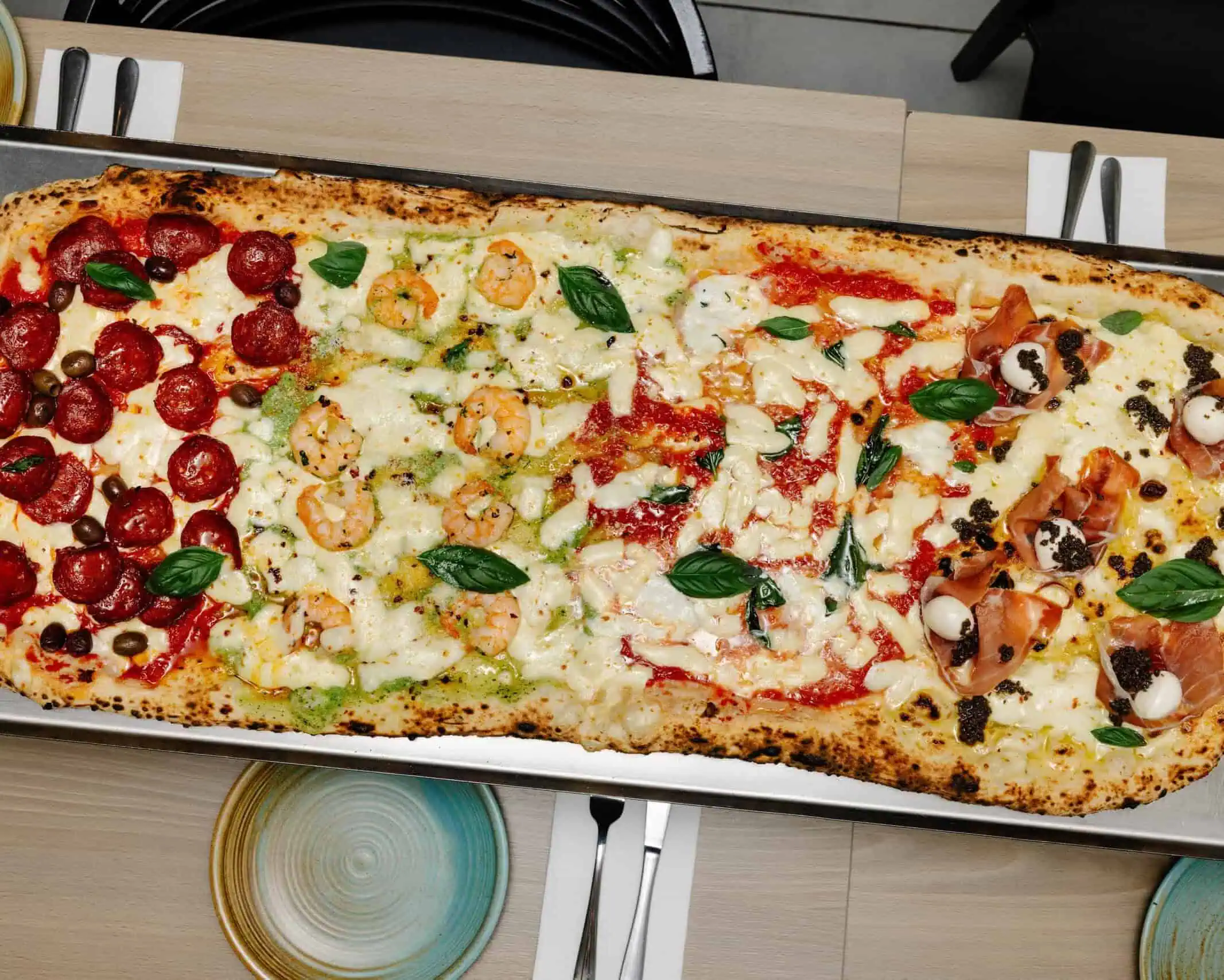 Extra-long metro pizza on a tray with four flavours—pepperoni with capers, prawn and pesto, tomato mozzarella basil, and prosciutto with truffle and mozzarella.