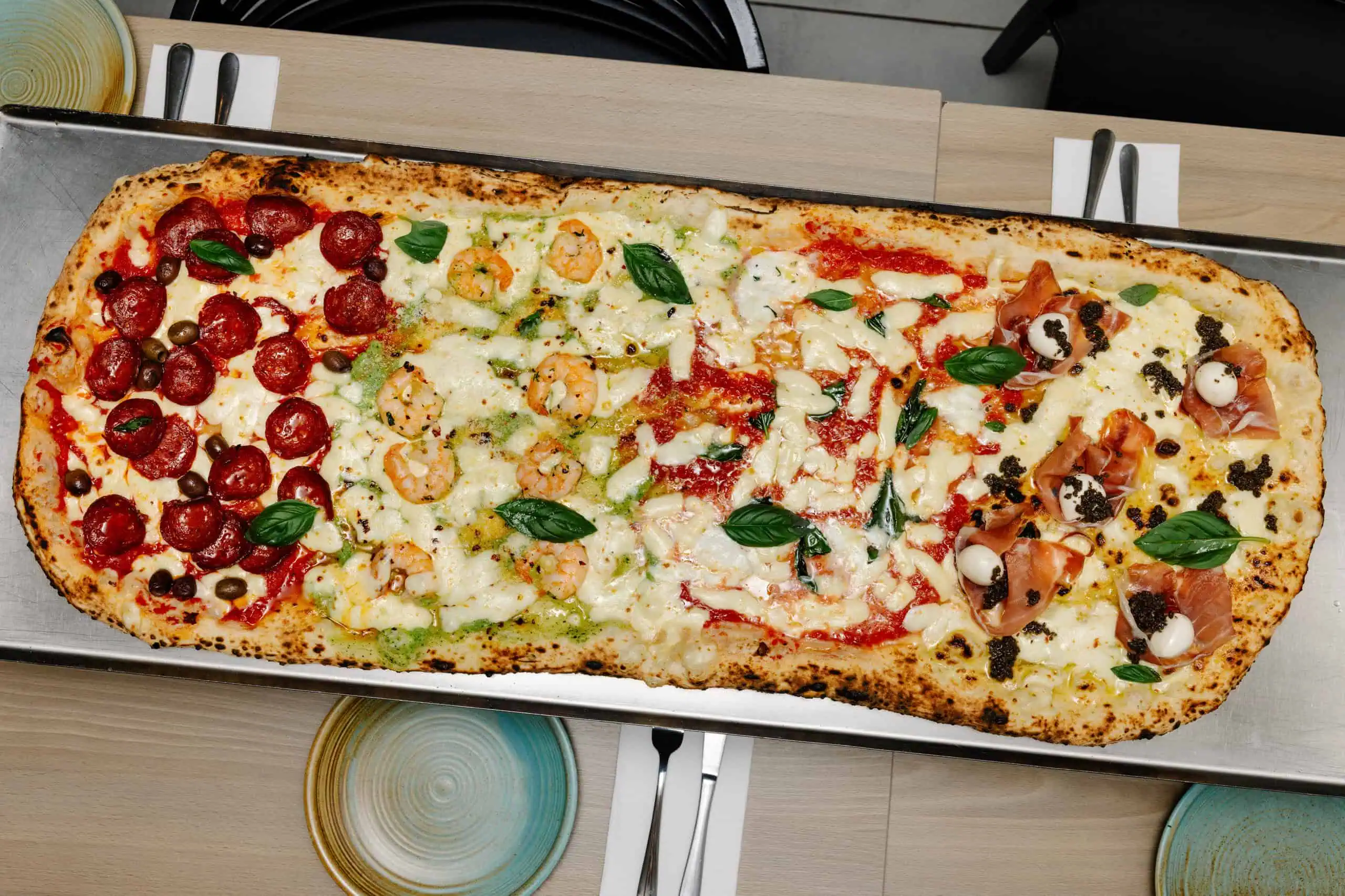 Extra-long metro pizza on a tray with four flavours—pepperoni with capers, prawn and pesto, tomato mozzarella basil, and prosciutto with truffle and mozzarella.