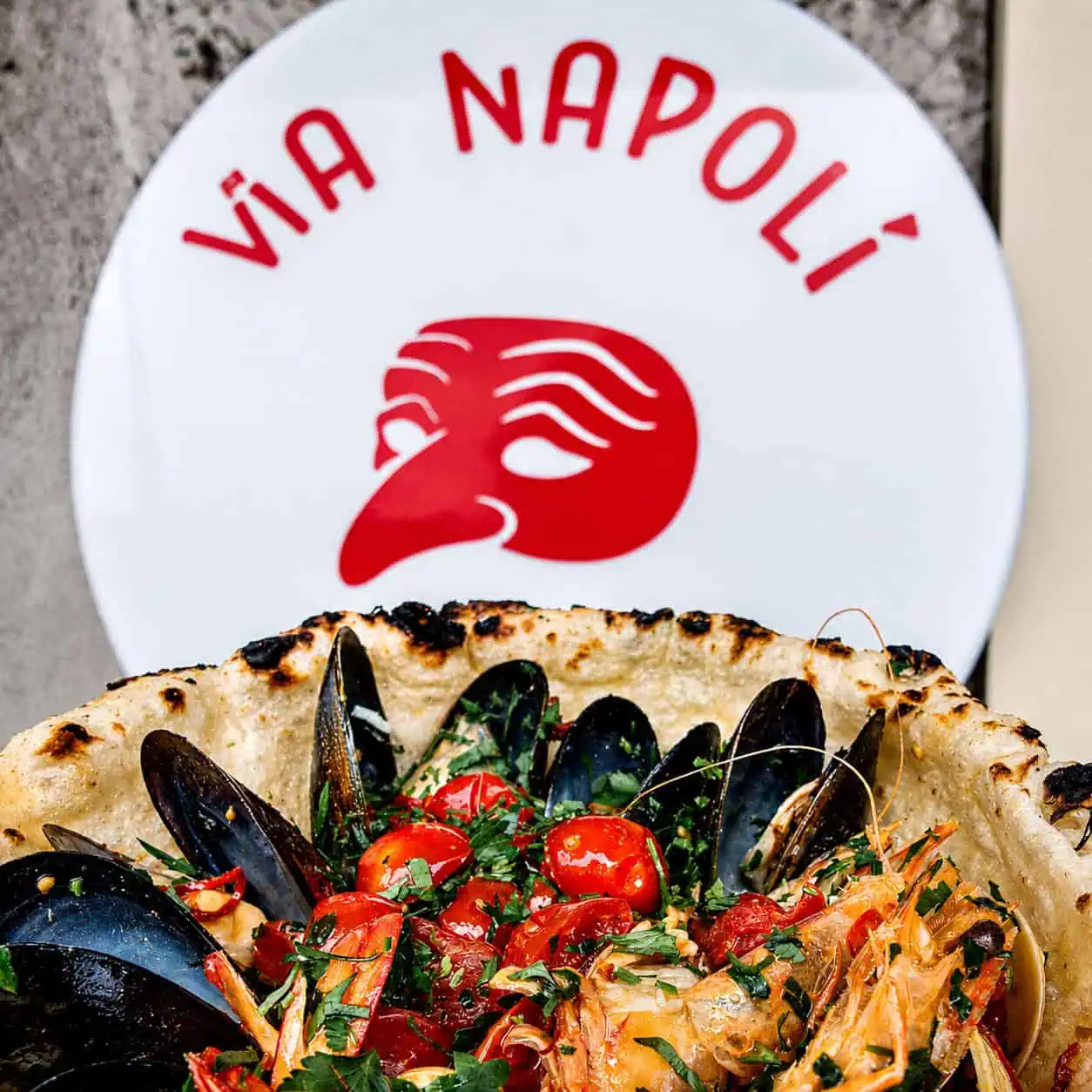 Close-up of Via Napoli seafood pizza topped with mussels, prawns, cherry tomatoes and fresh herbs, with Via Napoli branding in the background.