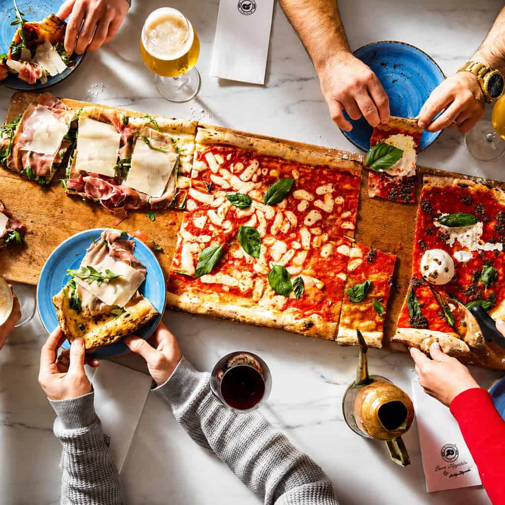 Overhead view of friends sharing a metre-long Via Napoli pizza with basil and drinks on a marble table
