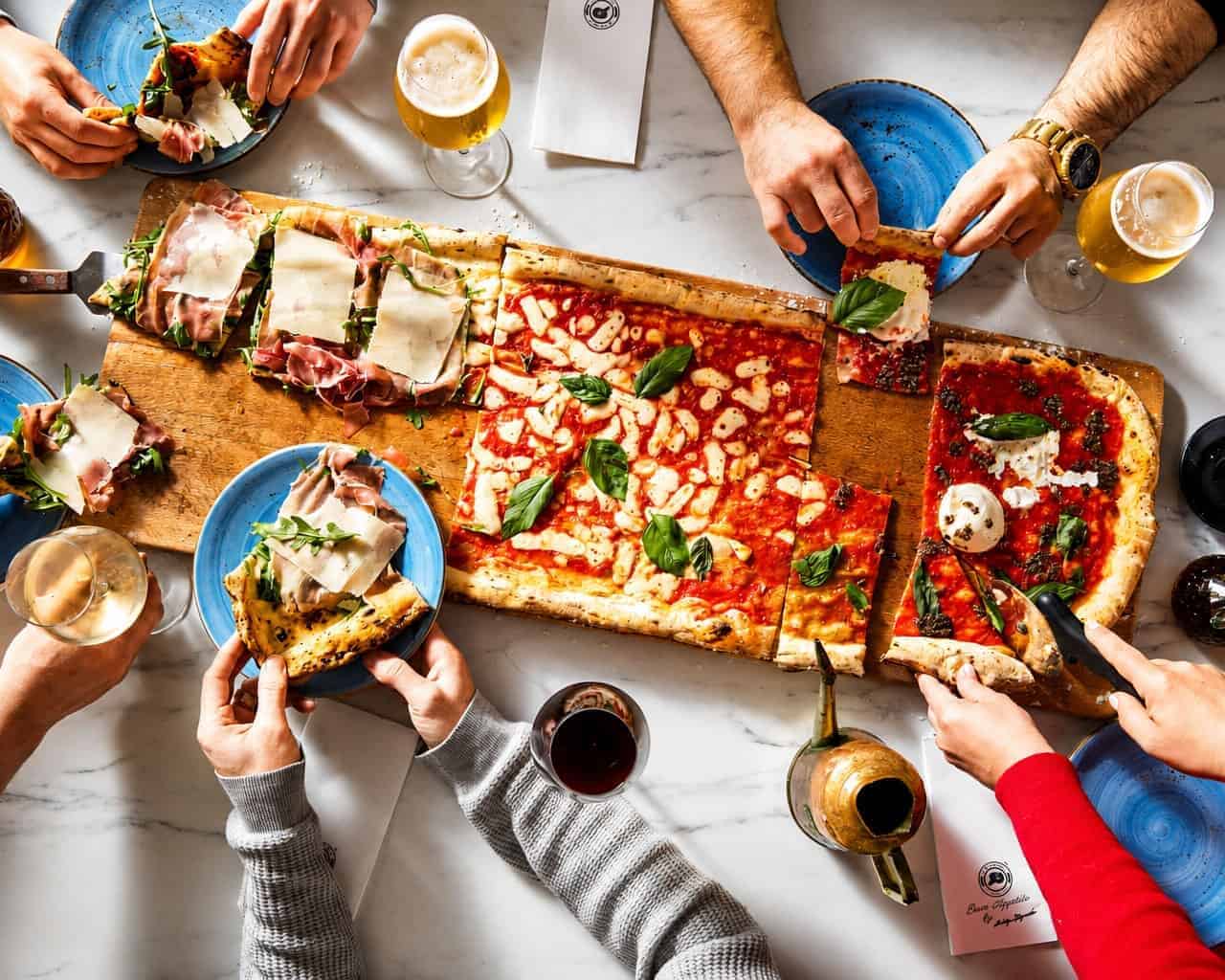 Overhead view of friends sharing a metre-long Via Napoli pizza with basil and drinks on a marble table