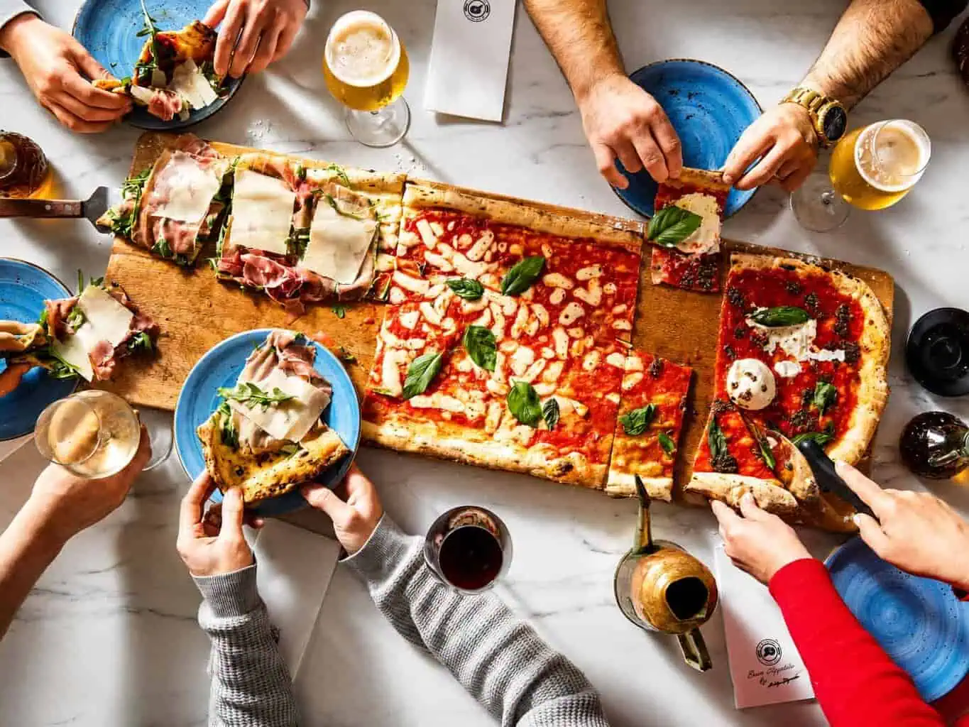 Overhead view of friends sharing a metre-long Via Napoli pizza with basil and drinks on a marble table