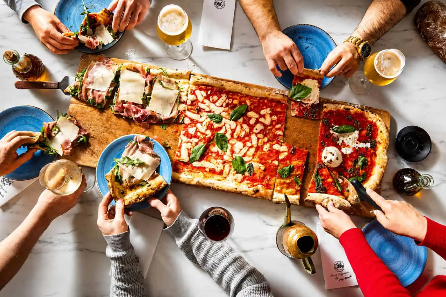 Overhead view of friends sharing a metre-long Via Napoli pizza with basil and drinks on a marble table