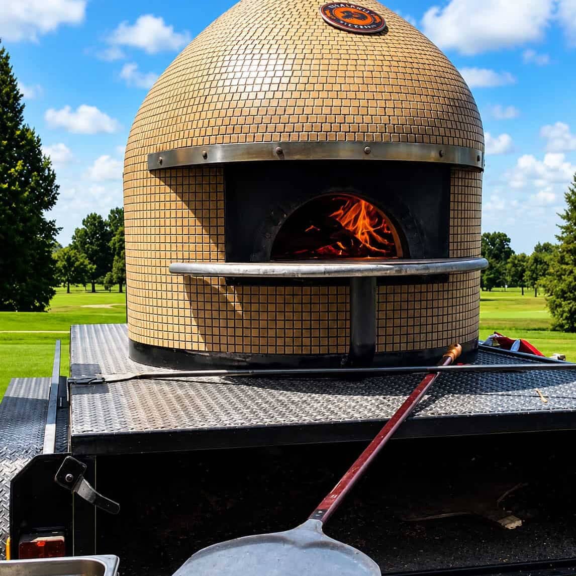 Mobile wood-fired pizza oven on a catering trailer with flames inside and a pizza peel in the foreground.