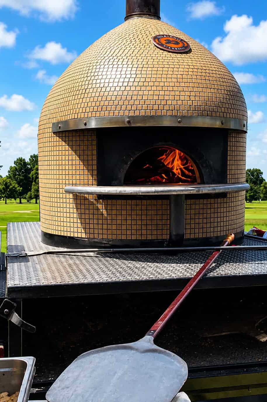 Mobile wood-fired pizza oven on a catering trailer with flames inside and a pizza peel in the foreground.