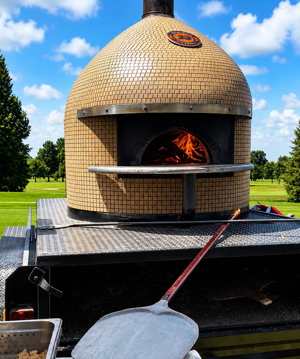 Mobile wood-fired pizza oven on a catering trailer with flames inside and a pizza peel in the foreground.