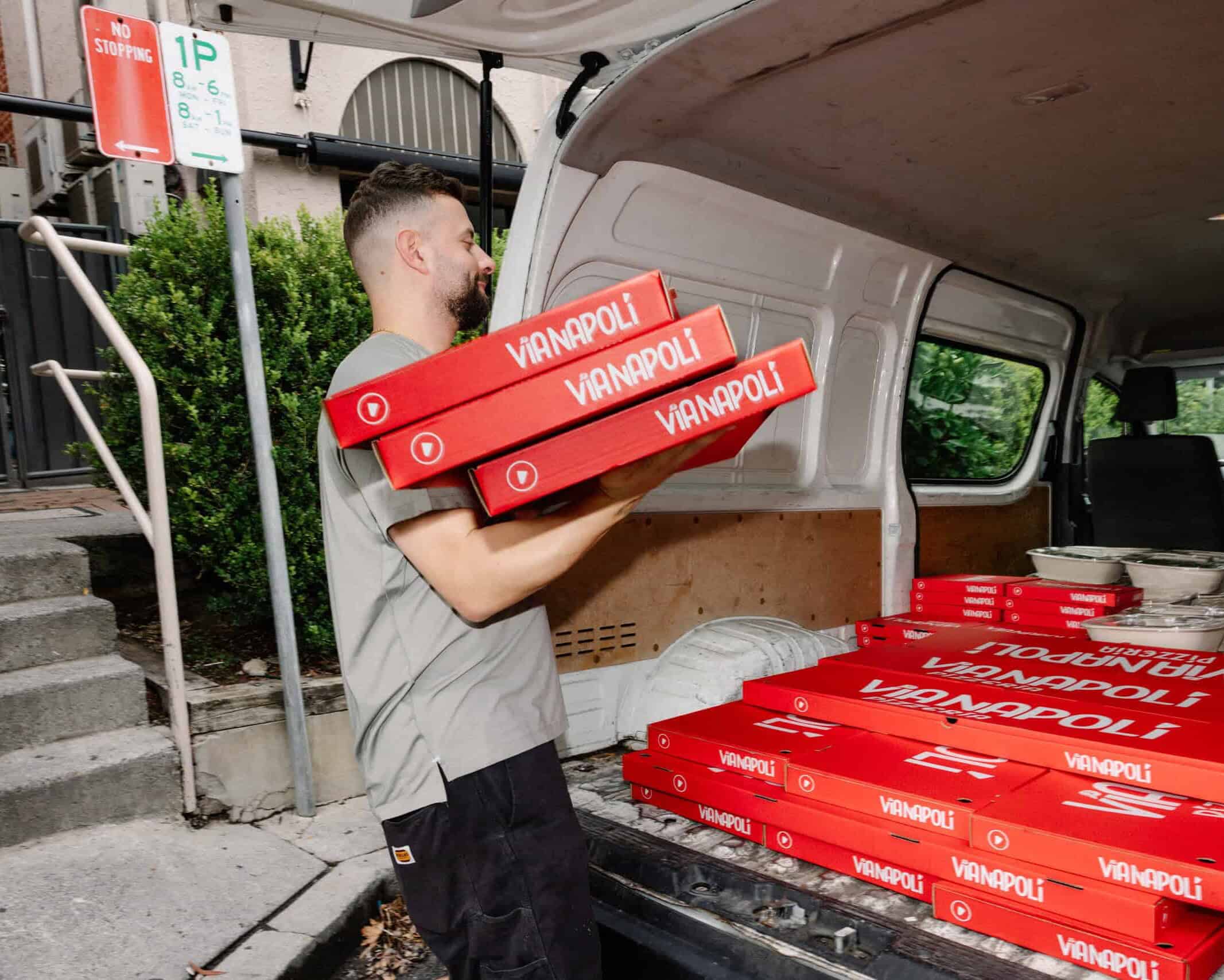 Staff member loading stacked red Via Napoli Pizzeria pizza boxes into a delivery van with catering meals in containers.