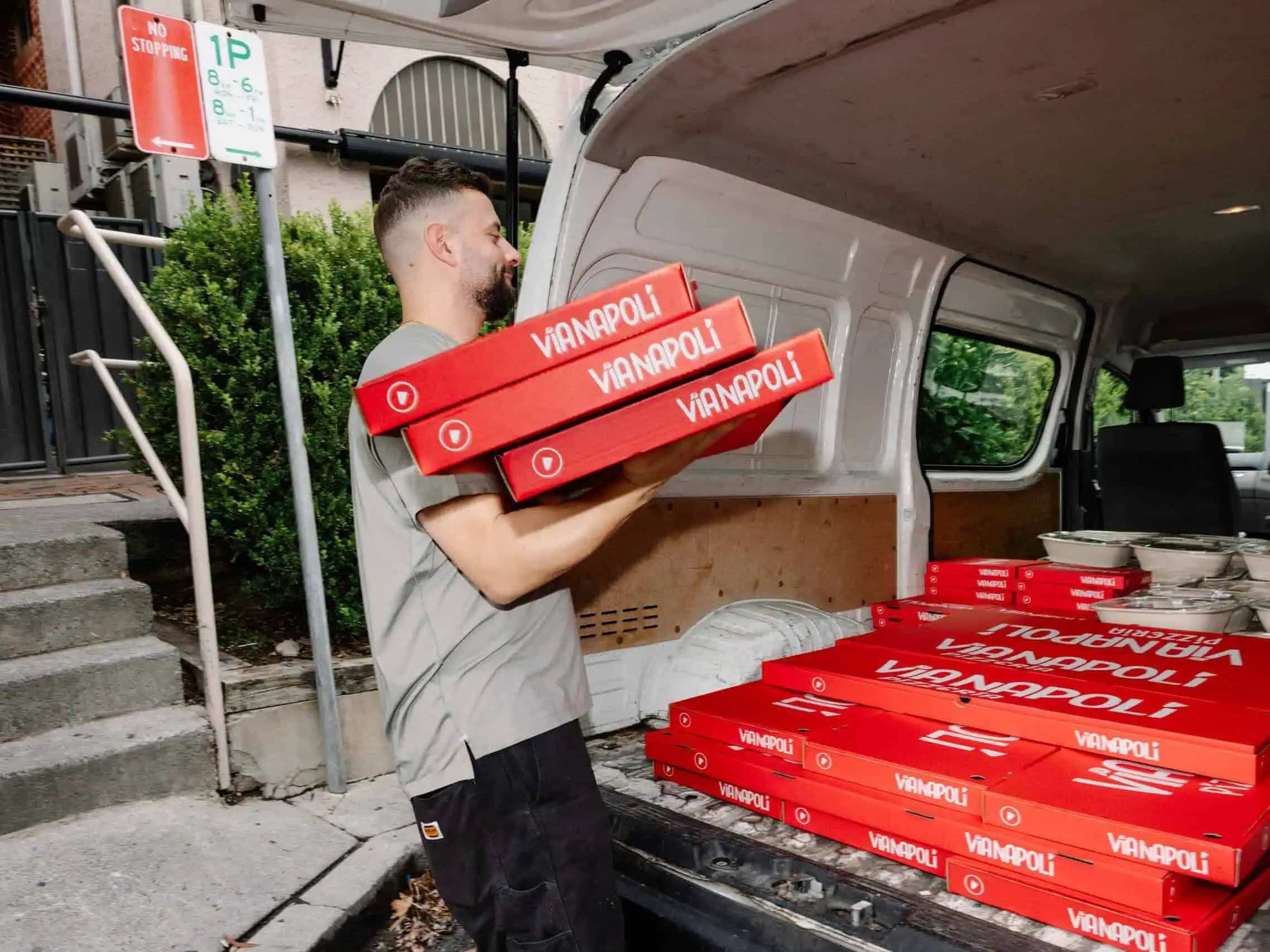 Staff member loading stacked red Via Napoli Pizzeria pizza boxes into a delivery van with catering meals in containers.