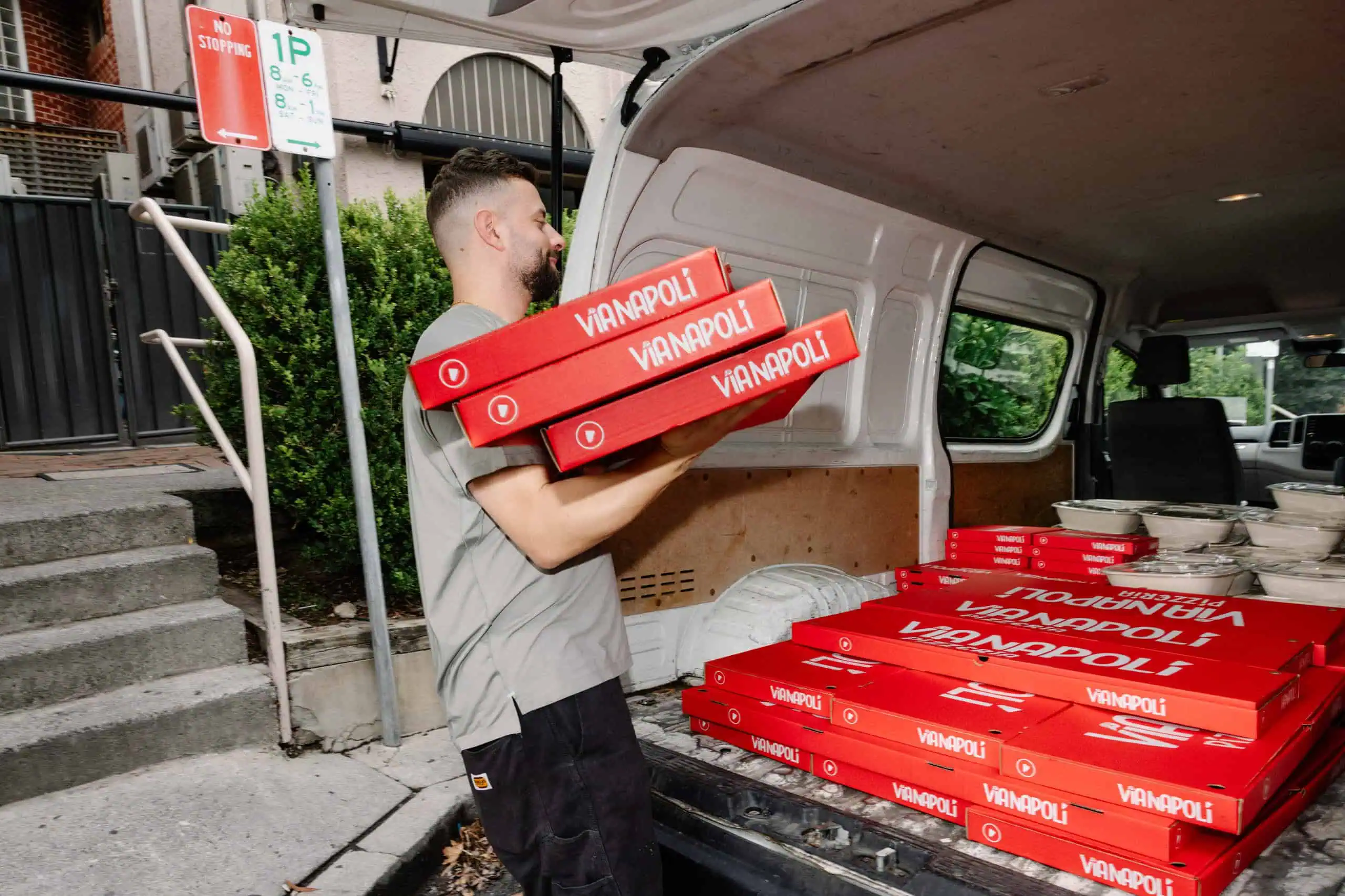 Staff member loading stacked red Via Napoli Pizzeria pizza boxes into a delivery van with catering meals in containers.