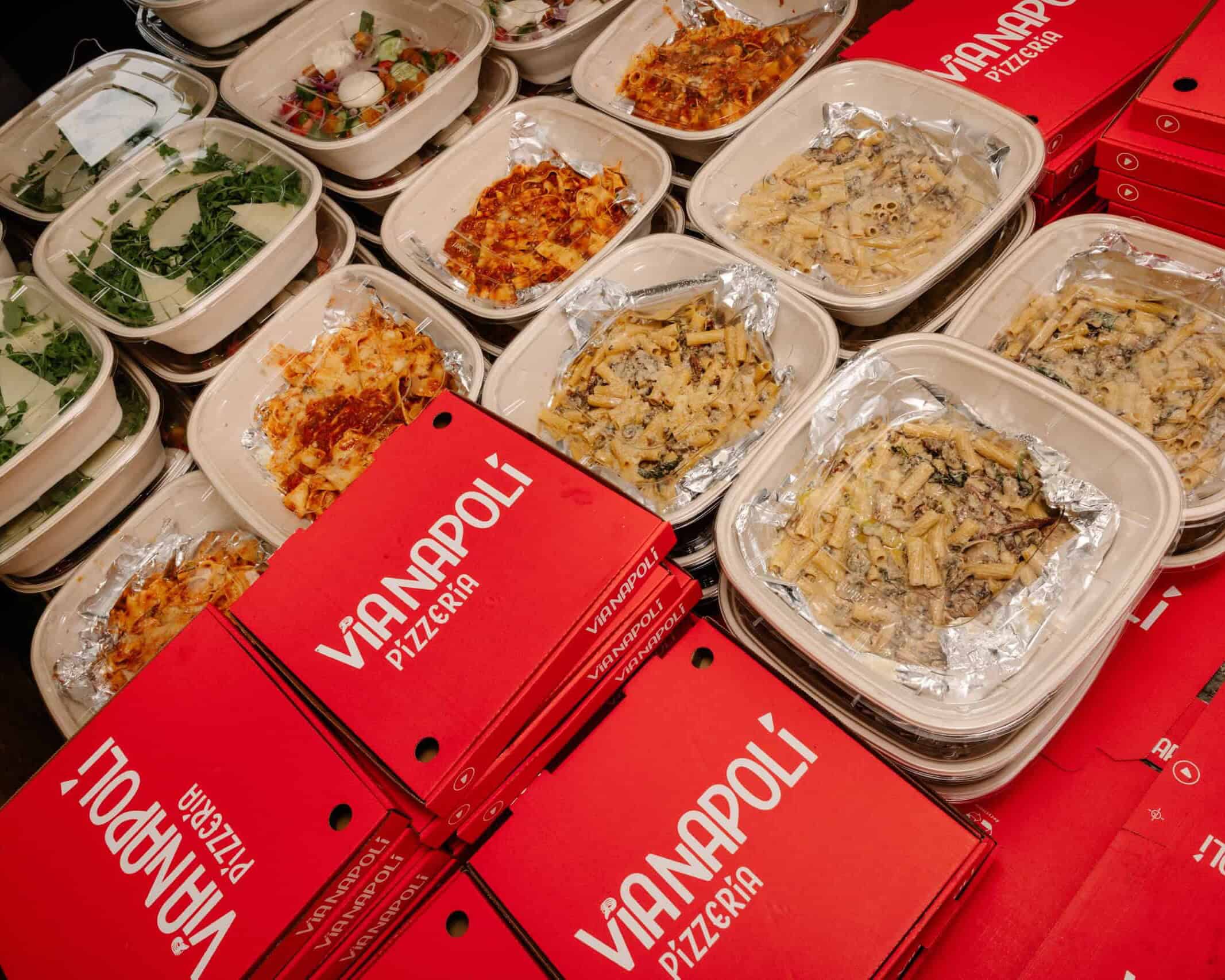 Takeaway meal containers with creamy rigatoni pasta, lasagna and fresh salads beside red Via Napoli Pizzeria pizza boxes.