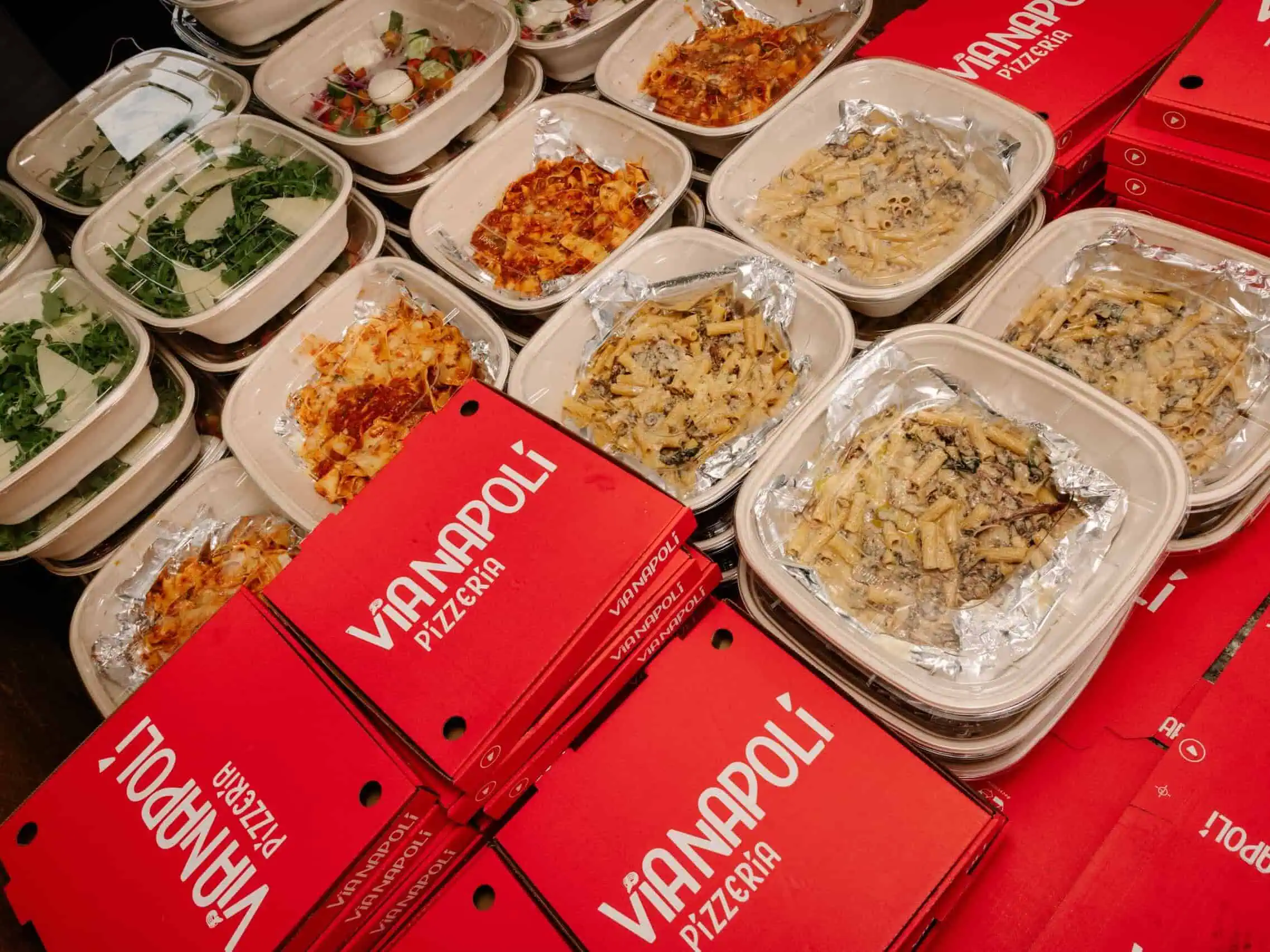 Takeaway meal containers with creamy rigatoni pasta, lasagna and fresh salads beside red Via Napoli Pizzeria pizza boxes.