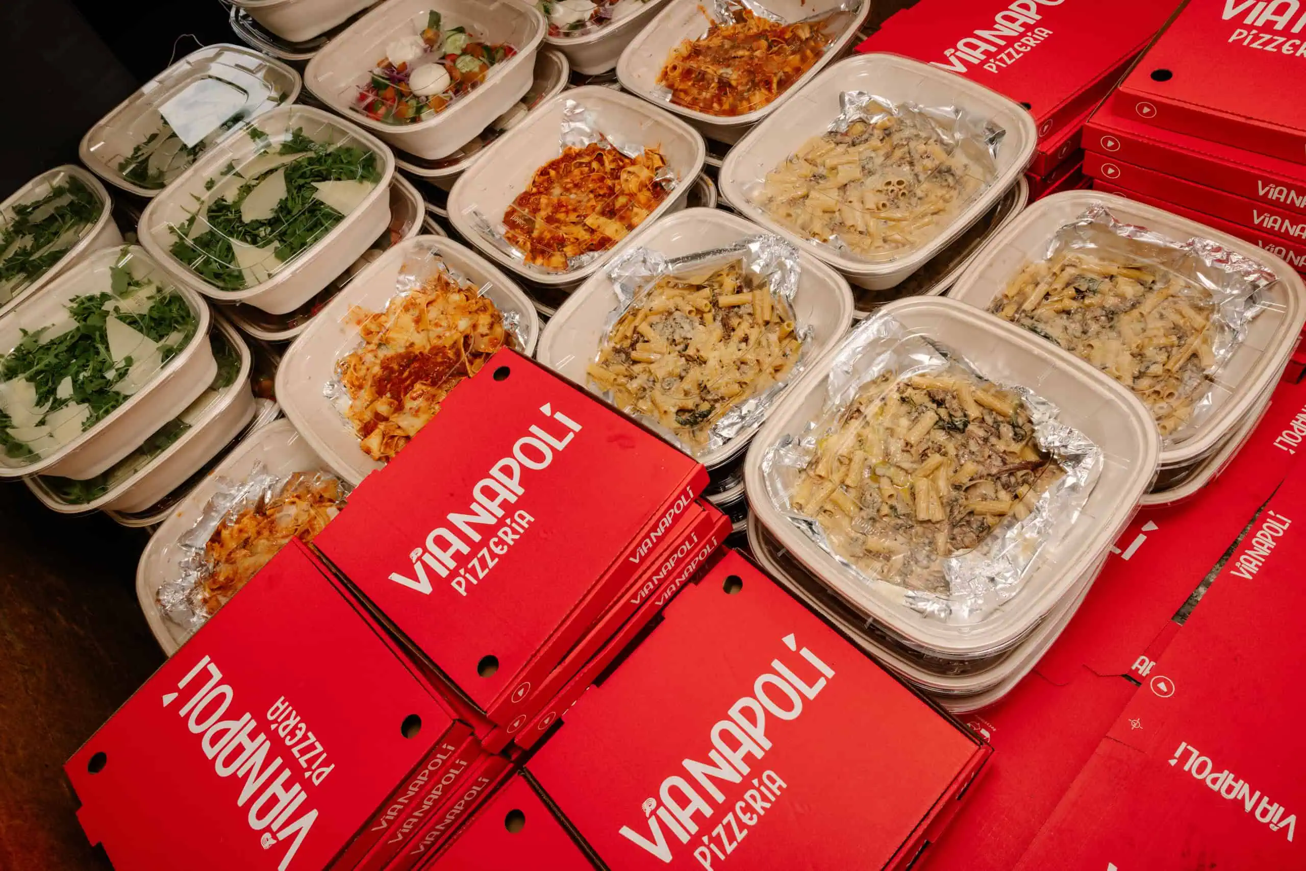 Takeaway meal containers with creamy rigatoni pasta, lasagna and fresh salads beside red Via Napoli Pizzeria pizza boxes.