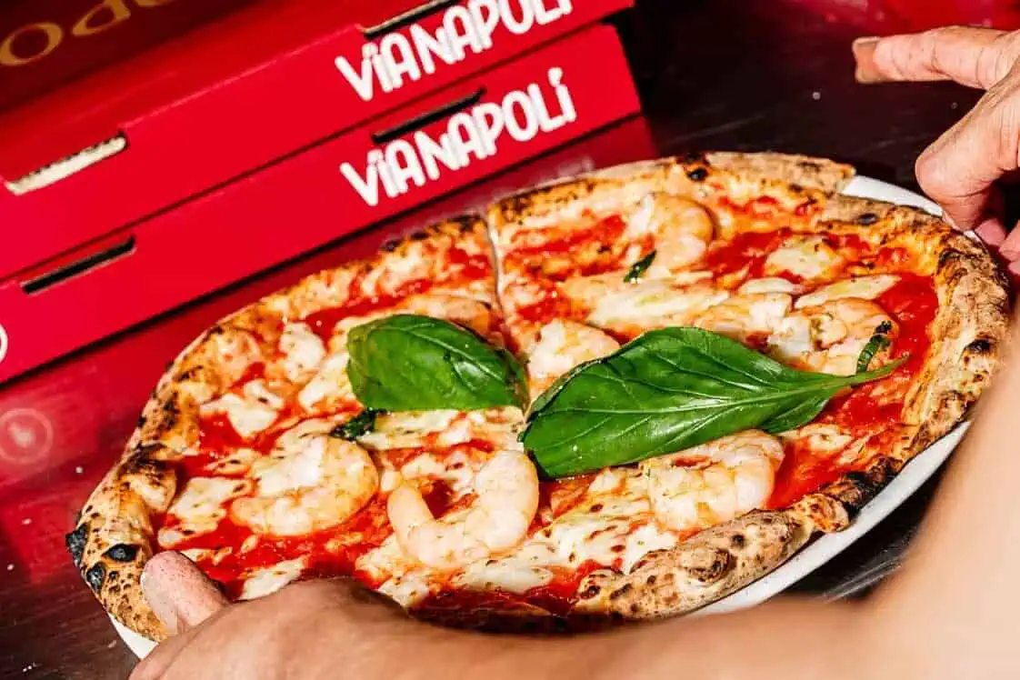 Hands holding a shrimp-topped Neapolitan pizza with fresh basil on a plate, with Via Napoli pizza boxes in the background.