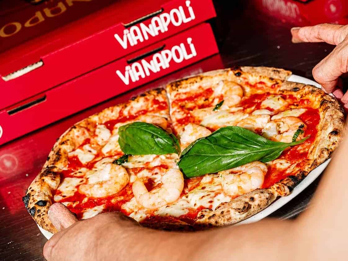 Hands holding a shrimp-topped Neapolitan pizza with fresh basil on a plate, with Via Napoli pizza boxes in the background.