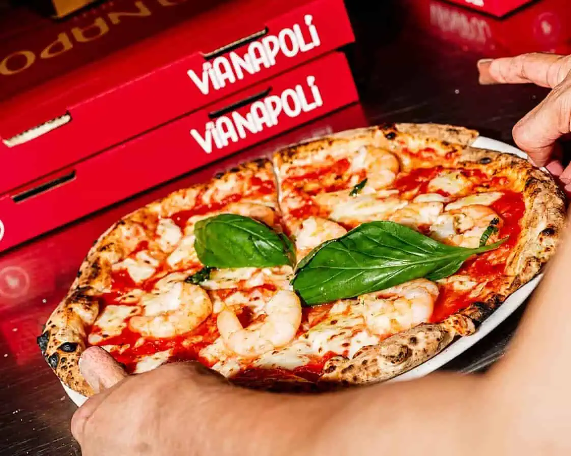 Hands holding a shrimp-topped Neapolitan pizza with fresh basil on a plate, with Via Napoli pizza boxes in the background.